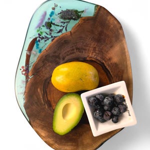 Puede incluir: Una tabla de servir de madera con borde natural, con una sección de resina azul claro con flores incrustadas. La tabla contiene un mango amarillo, un aguacate cortado por la mitad y un pequeño plato cuadrado de arándanos. El plato dice "SNACK TIME".