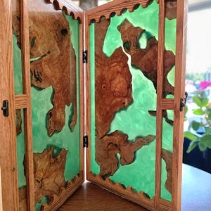 May include: A wooden backgammon board, open to reveal a vibrant green interior. The board features natural wood grain patterns set against the green resin. The frame is made of light-colored wood, with decorative cutouts along the top edge.