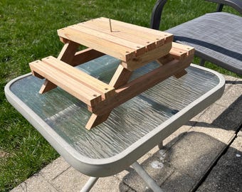 May include: A small wooden picnic table with a screw in the center of the tabletop. The table is sitting on a glass-topped table with metal legs.