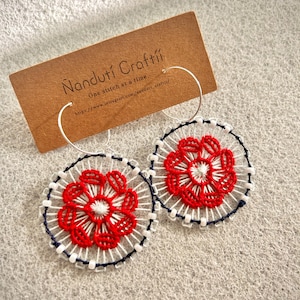 Peut inclure: Boucles d'oreilles créoles faites à la main avec un motif floral rouge, des accents blancs et bleu marine. Les boucles d'oreilles sont brodées et présentées sur un fond neutre. Diamètre d'environ 5 cm.