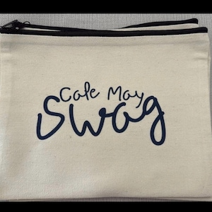 May include: Cream-colored canvas zippered pouch with the words "Cape May Swag" in navy blue script. The pouch has a black zipper and trim.