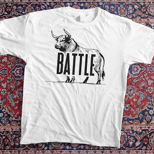 Peut inclure: T-shirt blanc avec un dessin noir d'un taureau et le mot "BATTLE" en lettres grasses. Le taureau est détaillé avec des cornes et une musculature. Le t-shirt est présenté sur un tapis à motifs.