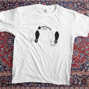 Peut inclure: T-shirt blanc avec un motif noir et blanc représentant une séquence de pas de danse. Le motif comprend des empreintes de pas, des flèches et les mots "Jump" et "Start". Le t-shirt est posé sur un tapis à motifs.