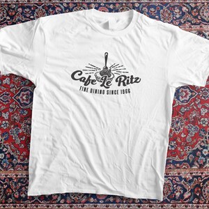 May include: White t-shirt with the text "Cafe Le Ritz Fine Dining Since 1966" in black. The design features a fork and a plate. The shirt is displayed on a patterned rug.
