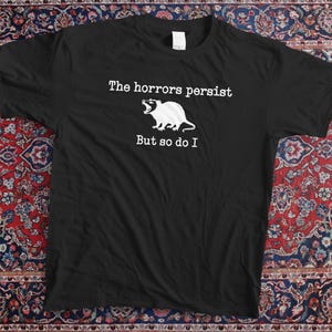 Peut inclure: T-shirt noir avec le texte "The horrors persist But so do I" et un dessin d'opossum blanc. Le t-shirt est posé sur un tapis à motifs.