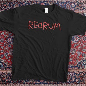 Puede incluir: Camiseta negra con la palabra "REDRUM" en rojo, con letras manuscritas. La camiseta está colocada sobre una alfombra estampada con colores rojo, azul y crema. La alfombra tiene un diseño intrincado.