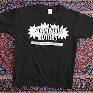 Peut inclure: T-shirt noir avec un motif en forme d'éclat d'étoile blanc portant l'inscription "Wormwood Motors" et "Quality Used Cars Since 1978". Le t-shirt est présenté sur un tapis à motifs.