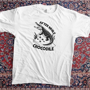 May include: White t-shirt featuring a black and white illustration of a crocodile with the text "AFTER WHILE CROCODILE". The design includes a detailed crocodile on a rock with grass.