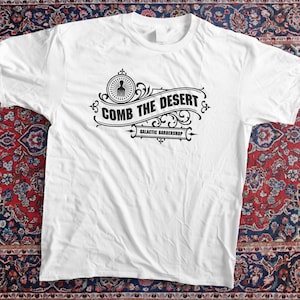 Peut inclure: T-shirt blanc avec un motif graphique noir. Le motif présente le texte "COMB THE DESERT" dans une bannière, avec "GALACTIC BARBERSHOP" en dessous. Le t-shirt est présenté sur un tapis à motifs.