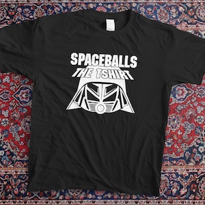 Peut inclure: T-shirt noir avec l'inscription blanche "SPACEBALLS THE TSHIRT" et un dessin blanc de casque. Le t-shirt est présenté sur un tapis à motifs.
