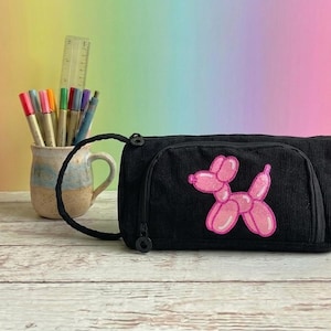 May include: Black corduroy pencil case with a pink balloon dog patch. The case has a zippered closure and a small handle. A ceramic mug with pens and a ruler are in the background.
