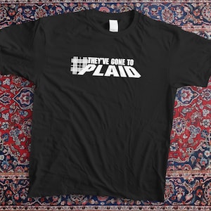 Peut inclure: T-shirt noir avec l'inscription blanche "THEY'VE GONE TO PLAID". Le t-shirt est présenté sur un tapis à motifs rouges, bleus et crème. Le t-shirt est à col rond et à manches courtes.