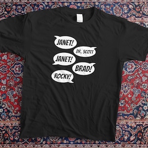 Peut inclure: T-shirt noir avec des bulles de texte blanches portant les noms "Janet", "Dr. Scott", "Brad" et "Rocky". Le t-shirt est posé sur un tapis à motifs aux couleurs rouge, bleu et crème.