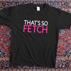 Może przedstawiać: Czarny t-shirt z napisem "THAT'S SO FETCH" w białym i różowym kolorze. Koszulka leży na wzorzystym dywanie w odcieniach czerwieni, błękitu i kremu. Codzienna, swobodna odzież.