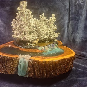 May include: A handcrafted wooden sculpture featuring a miniature landscape with a waterfall, pond, and silver-colored trees. The base is a cross-section of a tree trunk, with a dark blue background.