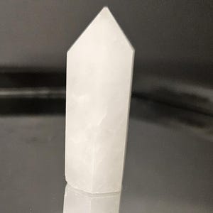 May include: A white, opaque, crystal point with a smooth, polished surface. The point is shaped like a pyramid with a flat base and a pointed top.