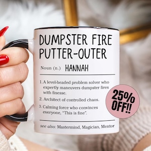 Op de afbeelding: Witte keramische mok met zwarte handgreep en rand, met de tekst "DUMPSTER FIRE PUTTER-OUTER" en de naam "HANNAH". De mok bevat een definitie en een "25% OFF!!" sticker.