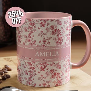 Puede incluir: Taza floral rosa y blanca con el nombre "AMELIA". La taza tiene un asa y borde rosa. Una pegatina con "25% OFF" es visible. La taza está sobre una superficie de madera.