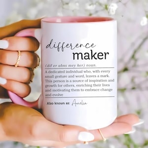 May include: White ceramic mug with a pink interior and handle, featuring the words "difference maker" in black script. The mug includes a definition and the name "Amelia" at the bottom. The mug is held by a person.