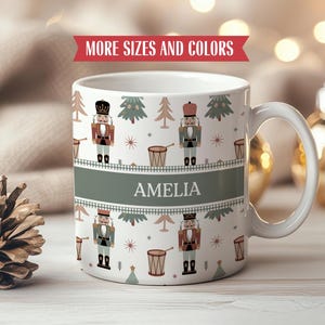 Pode incluir: Caneca de cerâmica branca com um padrão festivo de quebra-nozes e árvores de Natal. A caneca apresenta o nome "AMELIA" em uma faixa verde. As palavras "MORE SIZES AND COLORS" são exibidas em uma faixa vermelha.