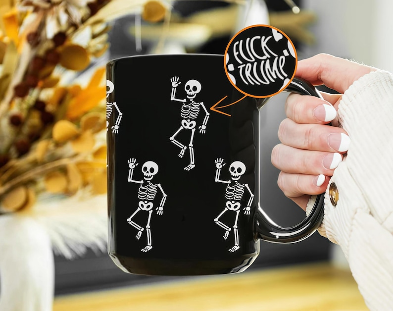 May include: Black ceramic mug featuring white skeleton illustrations. The mug has a black handle and the text "FUCK TRUMP" in a circle. The skeletons are waving. The mug is held by a person.