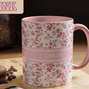May include: Pink floral-patterned mug with the name "CHLOE" in white letters. The mug has a pink interior and handle. The mug is decorated with a repeating pattern of red flowers and leaves on a white background.
