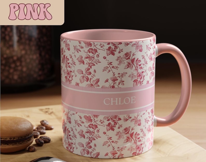 May include: Pink floral-patterned mug with the name "CHLOE" in white letters. The mug has a pink interior and handle. The mug is decorated with a repeating pattern of red flowers and leaves on a white background.