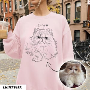 May include: Light pink sweatshirt featuring a black and white illustration of a Persian cat. The cat's name, "Lucy," is written above the image with a heart symbol. A smaller photo of the cat is in the lower right corner. The words "LIGHT PINK" are at the bottom.