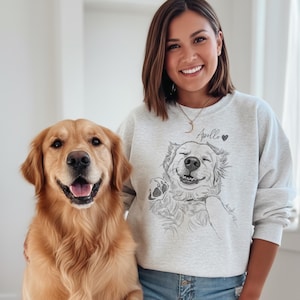 May include: A light gray sweatshirt featuring a smiling Golden Retriever illustration with the name "Apollo" and a heart. A real Golden Retriever dog is next to the person wearing the sweatshirt.