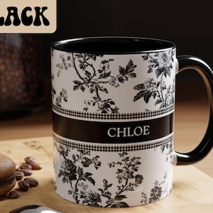 May include: A black and white floral-patterned mug with the name "CHLOE" printed on a black band. The mug has a black handle and interior. The word "BLACK" is in a stylized font in the upper left corner. A macaron and coffee beans are in the foreground.