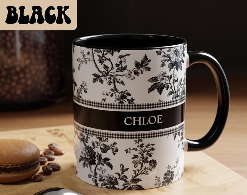 May include: A black and white floral-patterned mug with the name "CHLOE" printed on a black band. The mug has a black handle and interior. The word "BLACK" is in a stylized font in the upper left corner. A macaron and coffee beans are in the foreground.