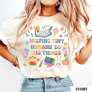 May include: Ivory t-shirt featuring the phrase "Helping Tiny Humans Do Big Things" in vibrant colours, accompanied by school-themed illustrations. The design includes the name "Mrs. Smith". The shirt is a light cream colour.