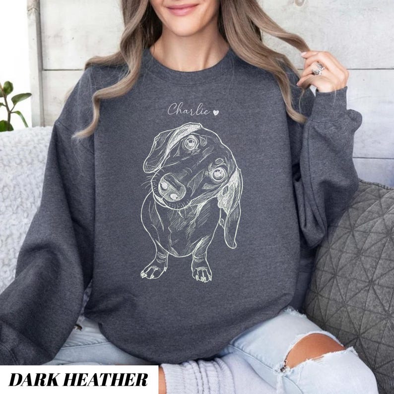 May include: Dark heather gray sweatshirt featuring a white line drawing of a dachshund dog. The dog's name, "Charlie", is written above the dog in cursive. The words "DARK HEATHER" are printed in a white, bold font at the bottom of the sweatshirt.