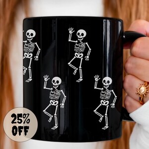 May include: A black ceramic mug featuring a repeating pattern of white skeleton illustrations. The skeletons are depicted in a waving pose. A circular sticker on the mug reads "25% OFF".