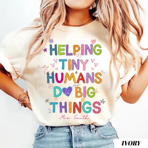 May include: Ivory-colored t-shirt with the text "HELPING TINY HUMANS DO BIG THINGS" in colorful, playful lettering. The design includes hearts, stars, and flowers. The name "Mrs. Smith" is written below the text.