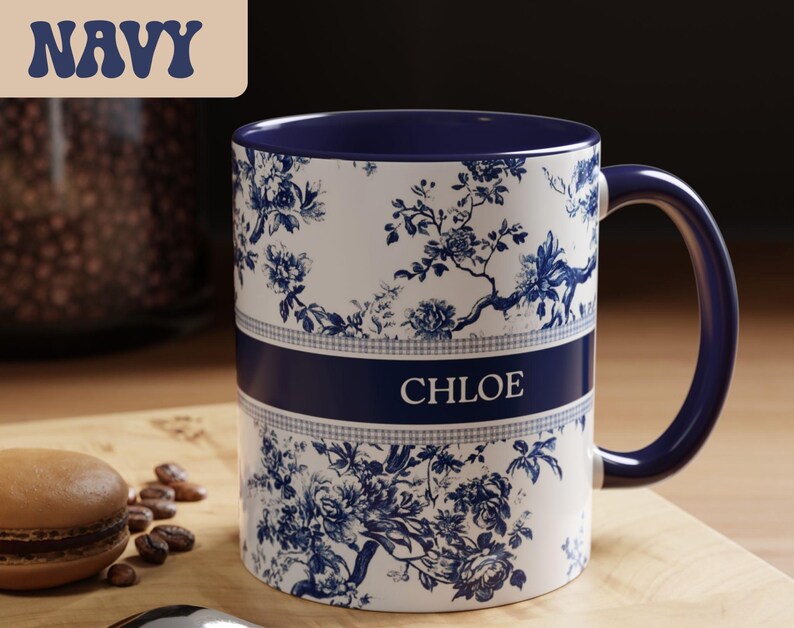 May include: A white ceramic mug with a navy blue handle and interior. The mug features a floral design in navy blue and the name "CHLOE" in white. The word "NAVY" is in a navy blue banner in the upper left corner.