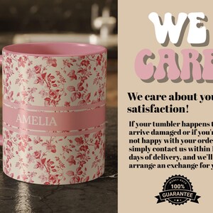 May include: A ceramic mug with a pink interior and handle, featuring a floral pattern in red and white. A pink band with the name "AMELIA" is wrapped around the mug. The mug is on a dark surface.