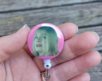 Könnte beinhalten: Eine rosafarbene, einziehbare Ausweishalterung mit einem kreisförmigen Design, das eine Frau mit blonden Haaren und einem überraschten Gesichtsausdruck zeigt. Die Halterung hat einen transparenten Kunststoffriemen und einen Metallclip.
