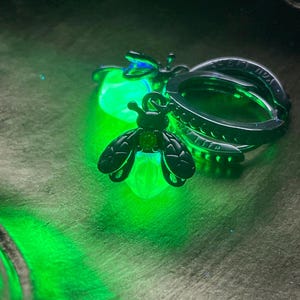May include: A dark green metal keychain with a stylized insect charm. The insect has a green gem and is attached to a ring with the words "YOU LIGHT UP MY LIFE" engraved on it. The insect charm glows with a bright green light.
