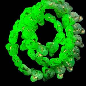 Könnte beinhalten: Eine Kette aus neongrünen Glasperlen in Form von Katzenköpfen, die jeweils goldfarbene Akzente an Ohren, Nase und Schnurrhaaren aufweisen. Die Perlen sind in einem kreisförmigen Muster vor einem schwarzen Hintergrund angeordnet.