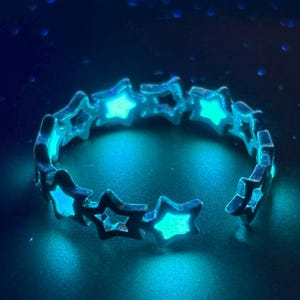 Può includere: Un anello argentato con una serie di ritagli a forma di stella. Ogni stella è riempita con un materiale luminoso di colore turchese, creando un contrasto sorprendente sullo sfondo scuro. Il design dell'anello è aperto, consentendo una vestibilità regolabile.