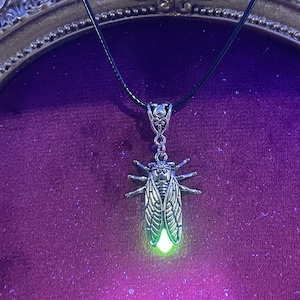 May include: A silver-toned pendant necklace featuring a detailed cicada design. The insect pendant has intricate wing and body details, with a glowing green light emanating from its abdomen. The necklace hangs on a black cord, suitable for everyday wear.