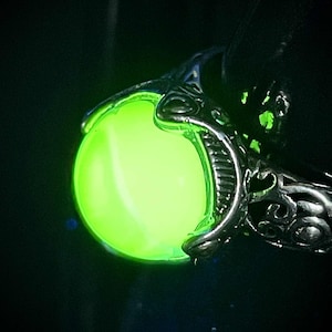 Puede incluir: Primer plano de un anillo plateado con un diseño ornamentado, que sostiene una piedra redonda verde lima brillante. El anillo se destaca sobre un fondo oscuro, resaltando el orbe luminoso.