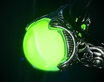 May include: A close-up of a silver ring with an ornate design, holding a glowing, round, lime-green stone. The ring is set against a dark background, highlighting the vibrant, luminous orb.