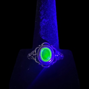 Könnte beinhalten: Ein silberner Ring mit einem ovalen, grünen Stein. Der Stein leuchtet unter UV-Licht hellgrün.