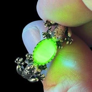 Może przedstawiać: Zbliżenie złotego pierścionka z żabą z świecącym zielonym owalnym kamieniem. Projekt pierścionka przedstawia dwie figurki żab trzymające kamień. Tło jest czarne.