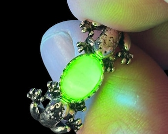 May include: A detailed close-up of a gold-colored frog ring with a glowing green oval stone. The ring's design features two frog figures holding the stone. The background is black.