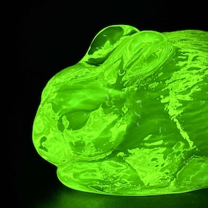 Op de afbeelding: Een neon-groen konijnbeeldhouwwerk tegen een zwarte achtergrond. Het beeldhouwwerk heeft een gladde, glanzende textuur en is verlicht, waardoor een gloeiend effect ontstaat. De vorm van het konijn is gedetailleerd, met zichtbare oren en gezichtskenmerken.