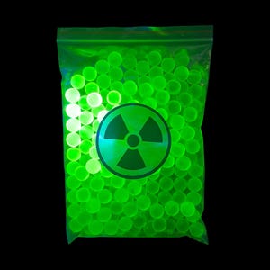 Puede incluir: Una bolsa de plástico transparente y resellable llena de numerosas esferas pequeñas, redondas y de color verde neón brillante. Un símbolo circular negro de peligro de radiación está impreso en la parte frontal de la bolsa sobre las cuentas.
