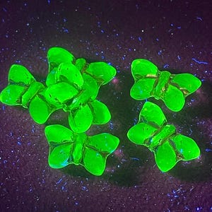 Puede incluir: Varios objetos brillantes verdes con forma de mariposa están dispuestos sobre una superficie púrpura oscura. Las mariposas tienen una apariencia translúcida, con detalles visibles en sus alas. El fondo es de un púrpura intenso con motas azules dispersas.